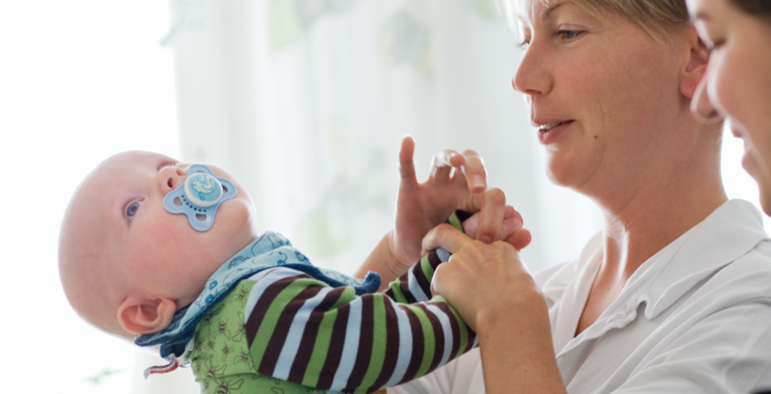 Barnmorska håller liten bebis i famnen. Bebisen har napp i munnen, lutar sig bakåt och tittar upp i taket.